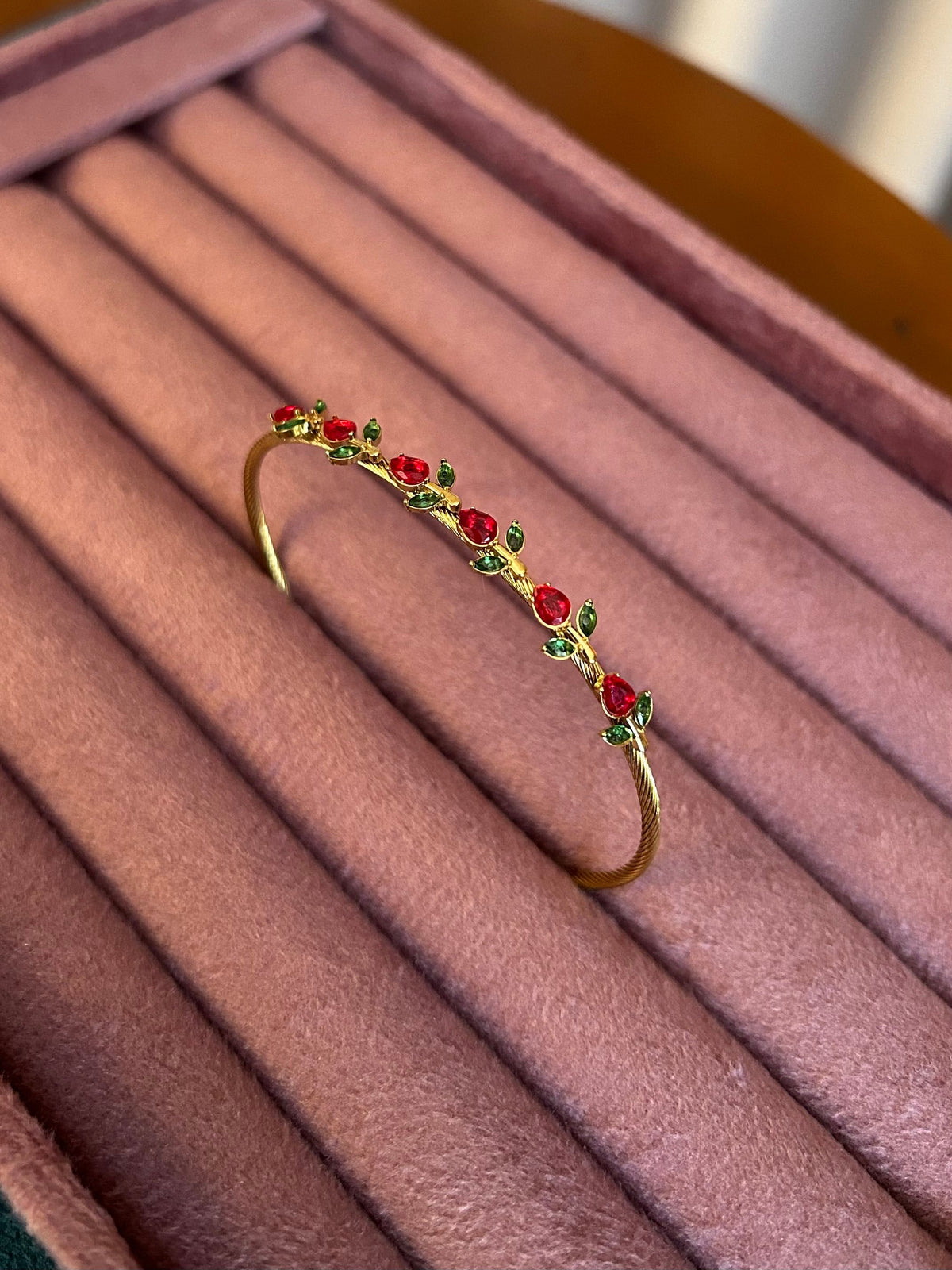 Gold twisted bangle with red crystal roses and green leaf accents on pink jewellery tray