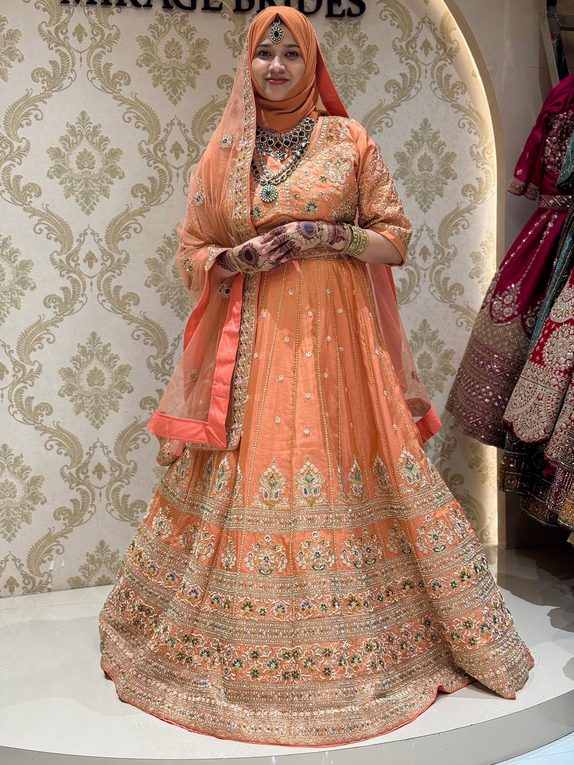 Woman in an ornate peach traditional outfit with a matching dupatta against a decorative wall.